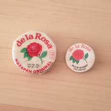 Load image into Gallery viewer, A real mazapan and a round button with a green and red floral design, labeled 'DE LA ROSA' and 'DULCED DE CACAHUETE' side by side for size comparison on a smooth light wood background.
